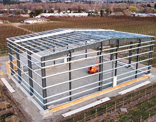 Aerial view of a steel frame building under construction in a rural setting, with the framework nearly complete. Visible are the sturdy girders and beams that form the skeleton of a large, industrial edifice. The surrounding area is dotted with greenery and farmland, underscoring the building's robust presence in a tranquil environment. Construction equipment is on site, indicating ongoing work and progress.