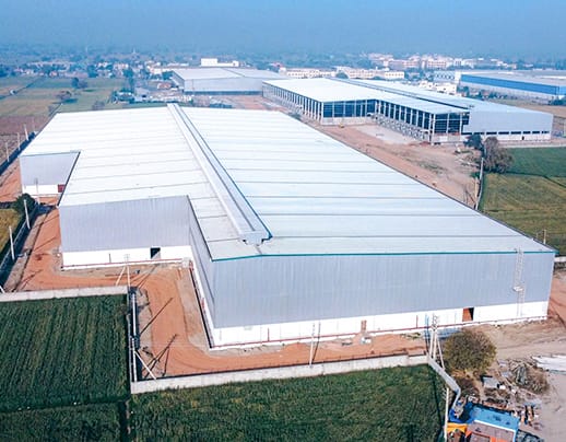 Aerial view of a sprawling metal workshop in an industrial zone, featuring a large, silver-colored corrugated metal roof and multiple loading docks. The facility is characterized by its modular steel construction and prefabricated components, designed for heavy-duty engineering and manufacturing activities.