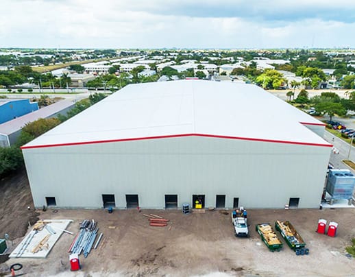 Drone view of a large prefab metal workshop with a prominent white facade and red trim, featuring a gabled roof design. The workshop's expansive structure is constructed with prefabricated steel panels, offering a modern, streamlined look. Materials and construction equipment are visible at the front, indicating active use and functionality.