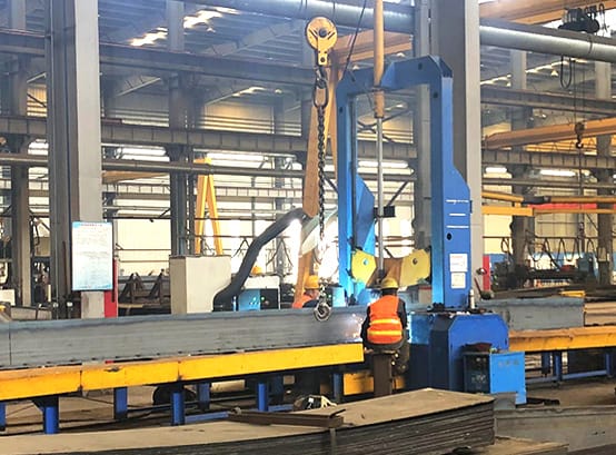In the midst of a steel structure fabrication workshop, a robust crane hoists a steel beam into place for welding. Surrounded by the essential equipment of the trade, the beam glows as it's expertly joined to form part of a durable framework, a testament to the skill involved in building strong, lasting structures.