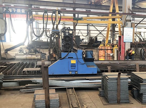 In a steel structure workshop, a powerful blue processing machine stands central, surrounded by organized stacks of steel bars and beams. The equipment is set for the day's work, signaling the start of a meticulous fabrication process.