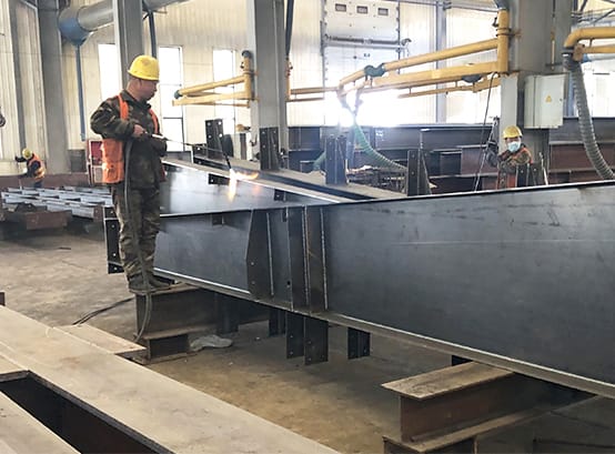 In the workshop of a steel manufacturing plant, a worker clad in safety equipment deftly handles a welding torch to join a sizable steel beam. Bright sparks from the torch illuminate the space where hefty girders are pieced together, forming the basis of substantial frameworks.