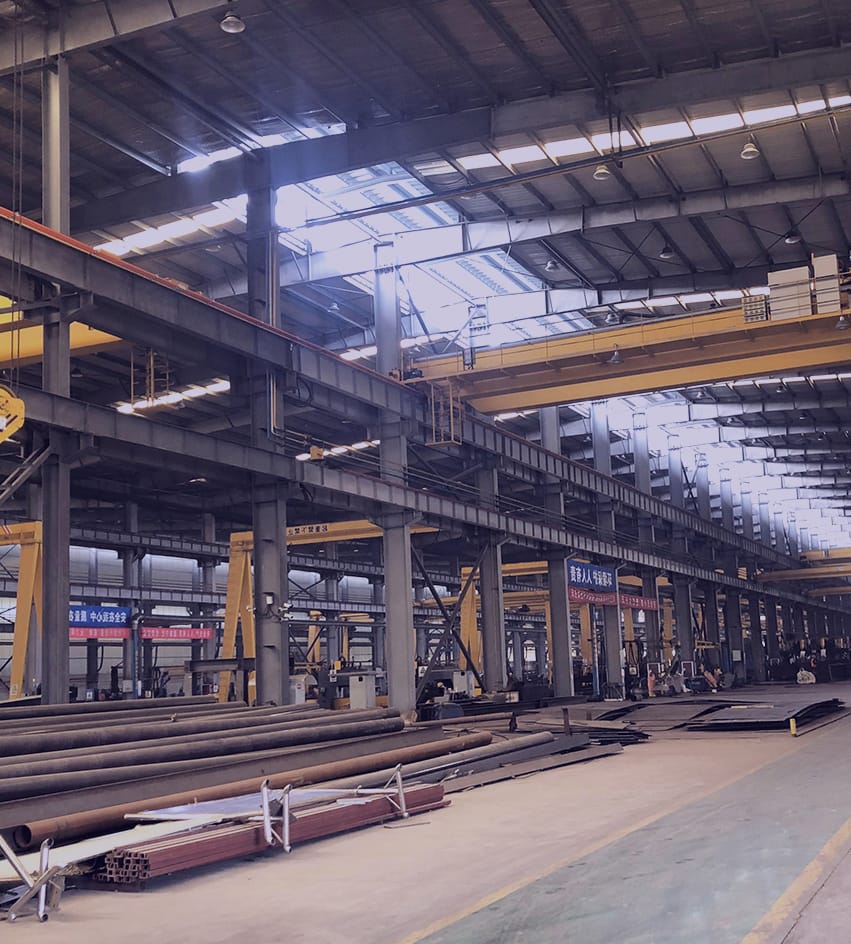 Interior of a structural steel supplier's production workshop, lined with heavy-duty steel beams and girders. The industrial space is equipped with overhead cranes and an array of machinery, indicative of large-scale steel fabrication capabilities. Stacks of steel pipes and metalwork materials are methodically arranged on the shop floor, ready for processing and distribution.